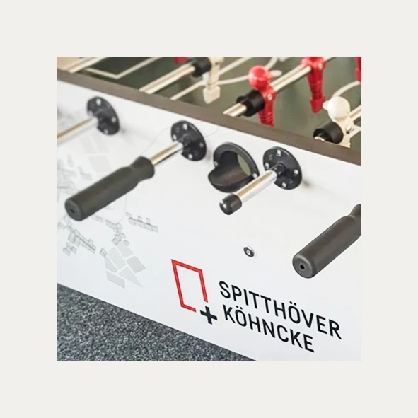 Kickertisch mit schwarzen und roten Spielstangen; auf der Seite ist das Logo und der Schriftzug 'Spitthöver + Köhncke' zu sehen.