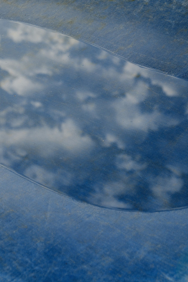 Auf einer blauen LKW Plane hat sich eine Pfütze gebildet. Wie in einem Spiegel spiegeln sich Wolken und der blaue Himmel in ihr.