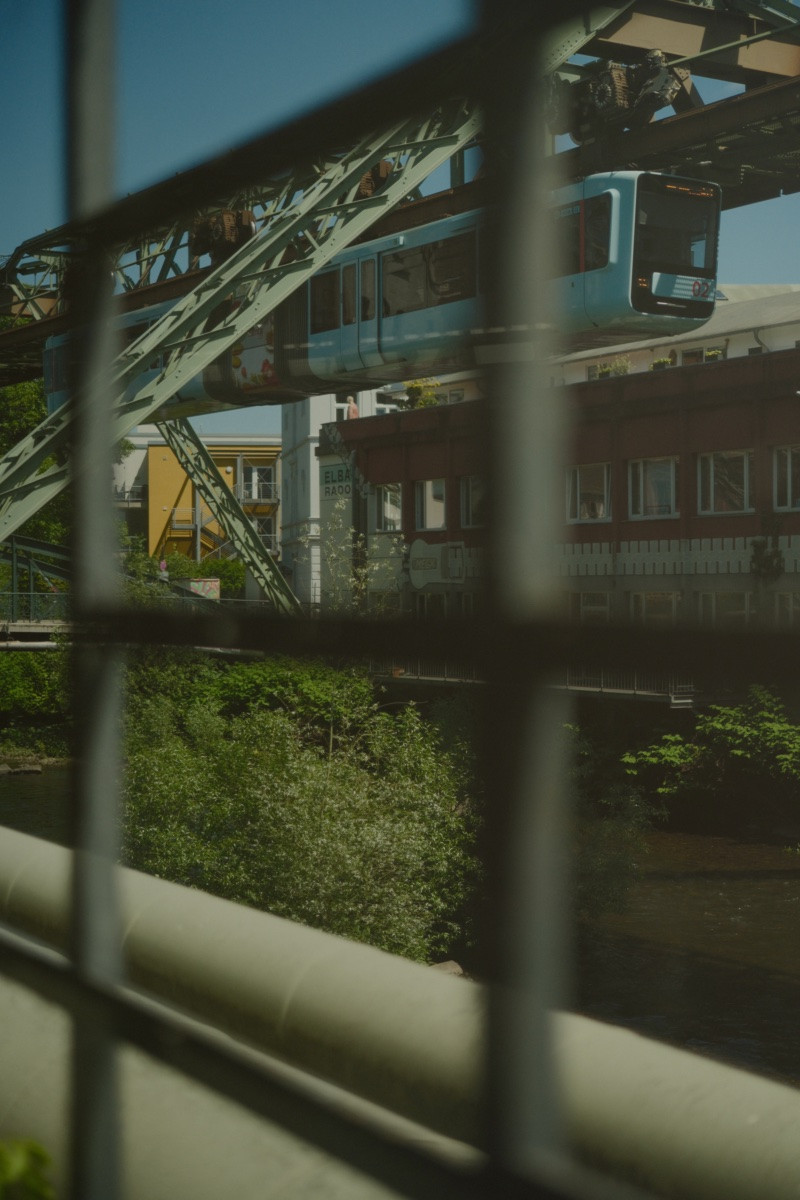 Blick durch ein unscharfes Sprossenfenster auf den Fluss Wupper. Am anderen Ufer reihen sich alte Industrie Gebäude auf. Über dem Fluss ist eine grüne Stahlkonstruktion gebaut unter der Mittig parallel zum Fluss eine hellblaue Schwebebahn entlang fährt.