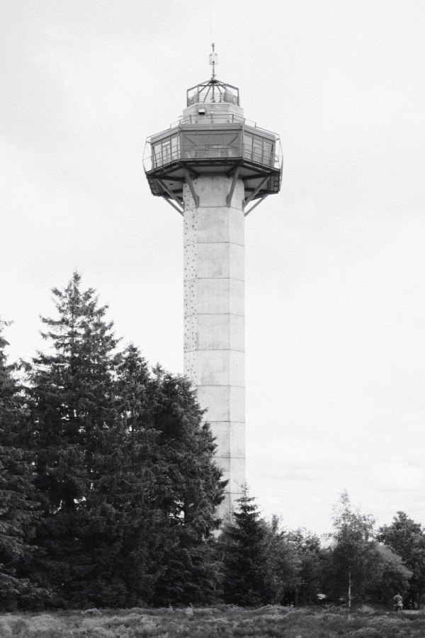 Schwarz-weiß Foto. Mittig im Bild ist ein hoher Turm aus hellem Beton vor grauem Himmel zu sehen. Eine Aussichtsplattform umläuft den Turm unter seiner Spitze. 

Vor dem Turm ist Heidelandschaft zu erkennen.