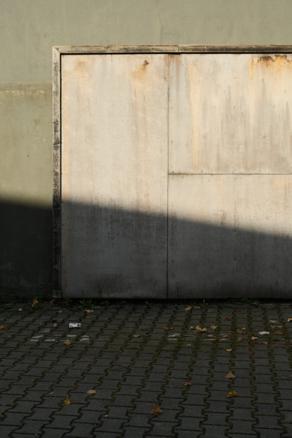 An einer grau-grünen Wand hängt eine leere aus Holzplatten gefertigte Plakatwand. Quer über die Wand verläuft ein dunkler Schatten.