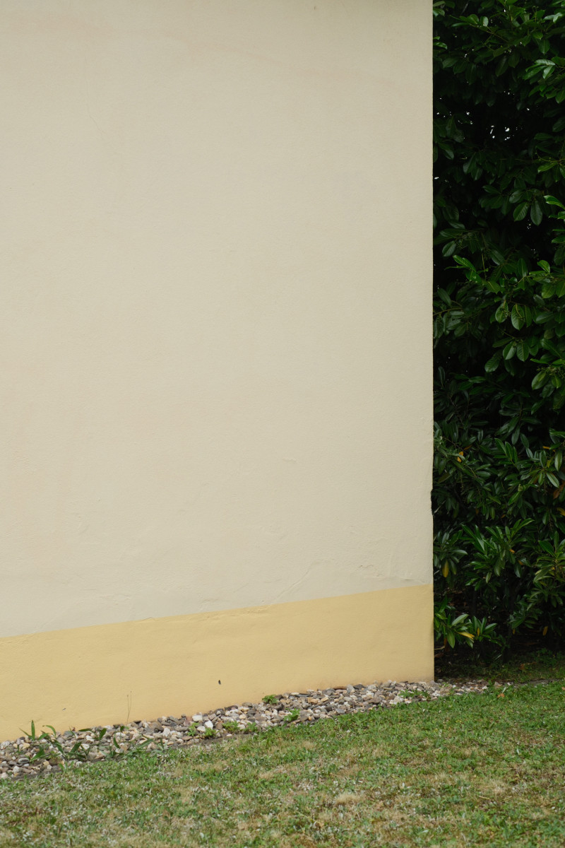 Eine gelb-beige Hauswand. Zum Boden ist sie mit einem 50cm hohen gelben Streifen abgesetzt. An der Hauswand entlang verläuft ein schmaler Kiesstreifen bevor dieser in eine kurz gemähte Rasenfläche übergeht. Rechts von der Hauswand wird das Bild in der vollen Höhe von dunkelgrünen Büschen ausgefüllt.