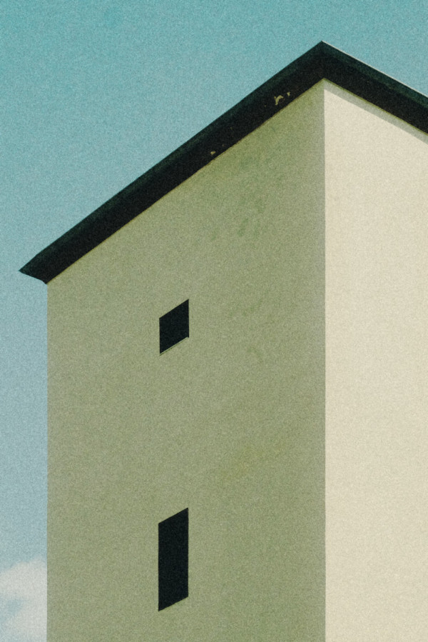 Foto eines hohen hell verputzten Gebäude-Turms. Zwei schmale schwarze Fenster sitzen übereinander mittig in der Wand. Sie wirken auf der hellen Fläche wie rechtwinklige Löcher.